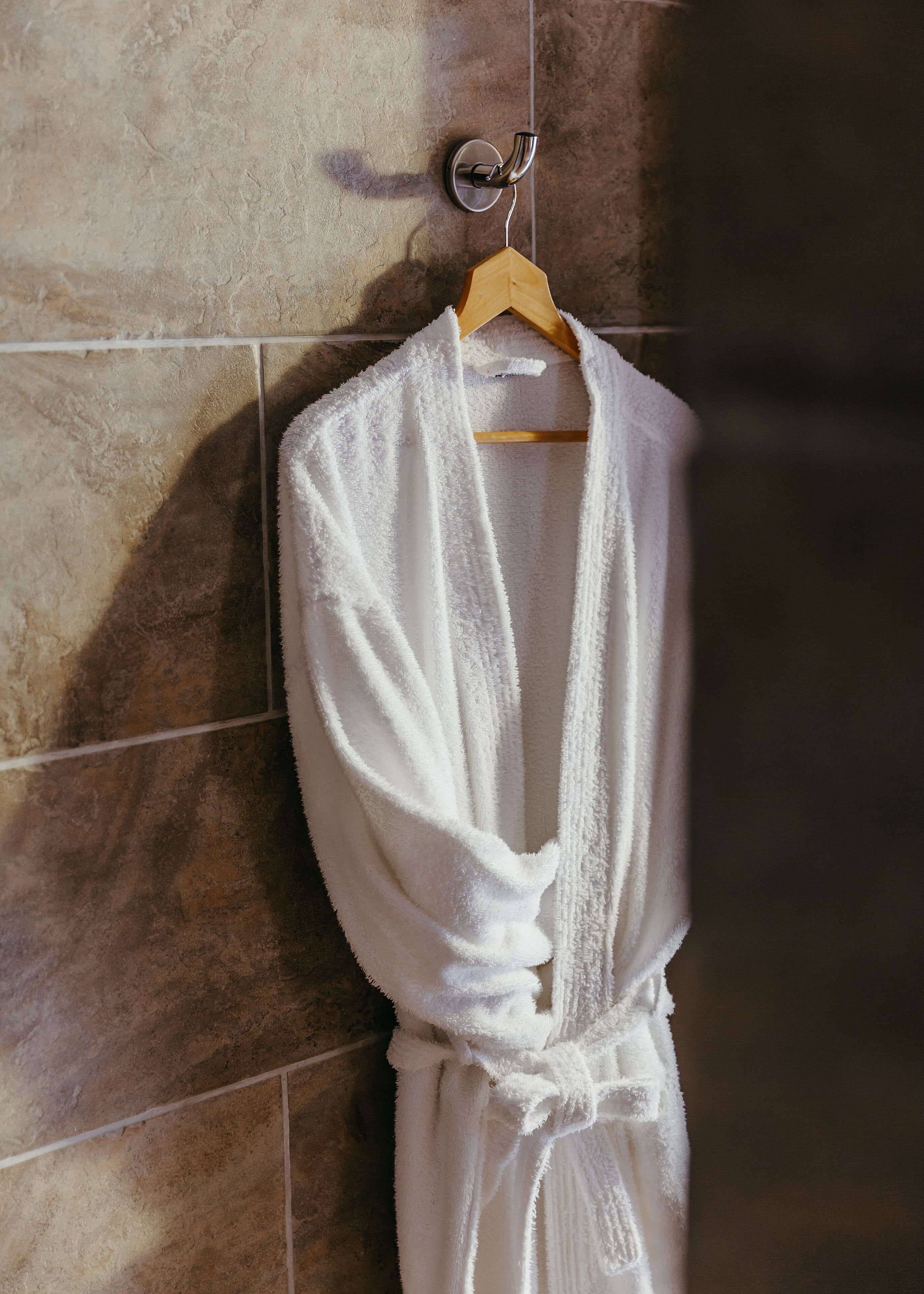 A white bathrobe hanging on a wooden hanger against a tiled wall.