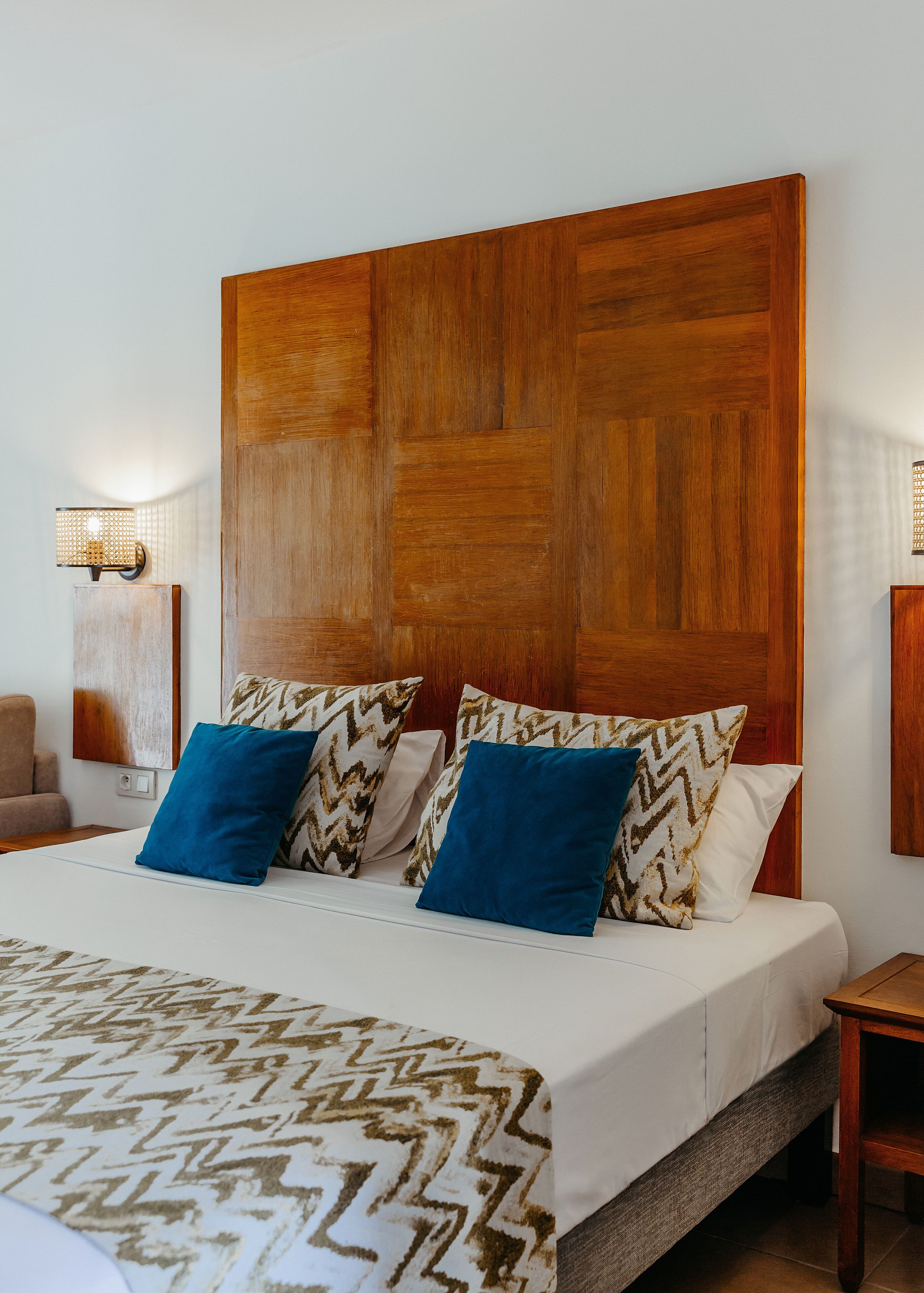 A hotel room with a large wooden headboard, a bed with patterned bedding, two blue pillows, and a bedside table with a lamp.