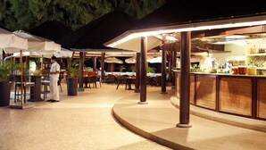 An outdoor dining area with tables and chairs under umbrellas, a bar with bottles and glasses, and a chef standing by the counter. - LUX* Saint Gilles (Saint-Paul)