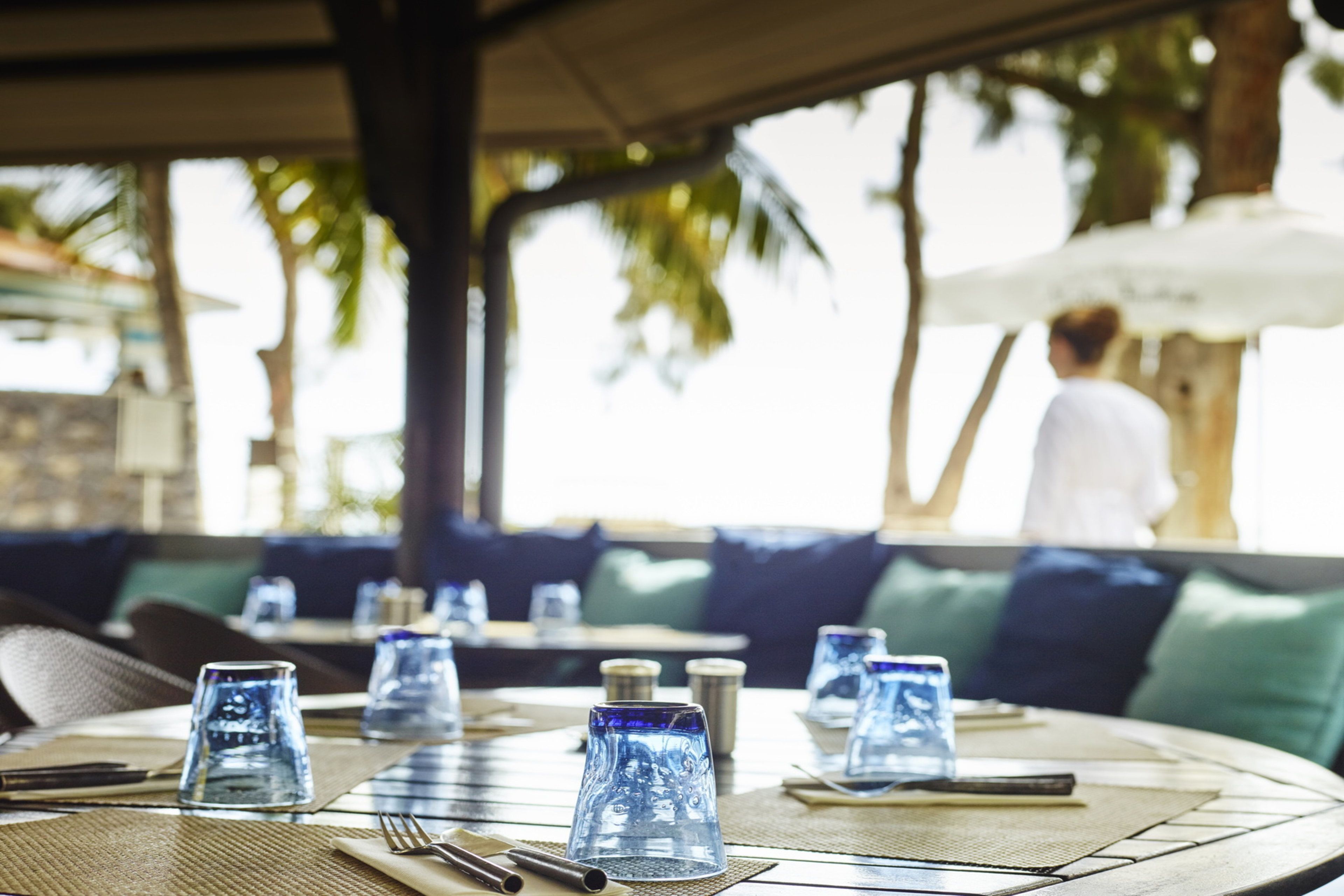 A dining area with tables set with blue glass tumblers, plates, and cutlery. There are green cushioned seating areas and a person in the background.