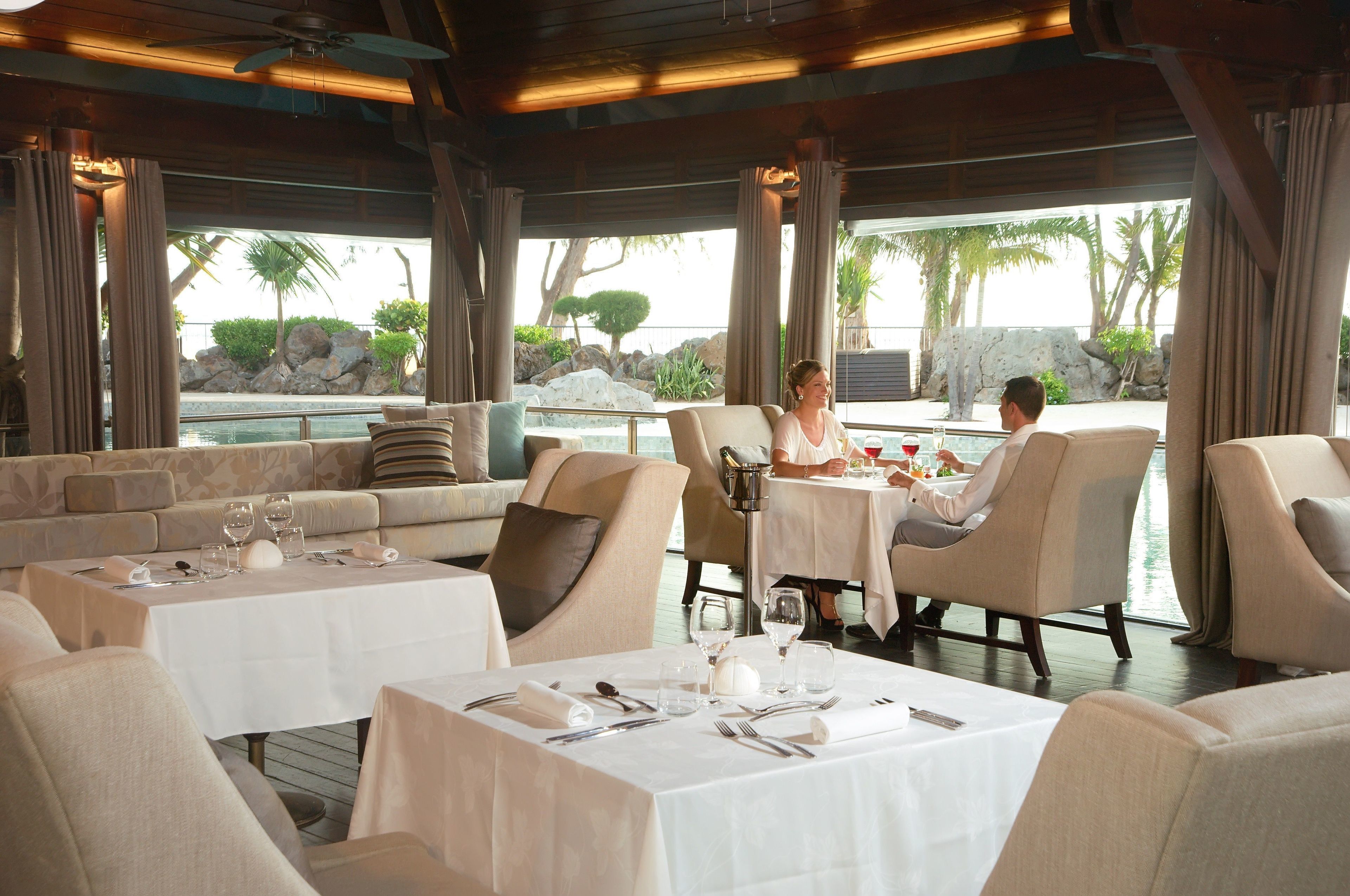 Ein Restaurant mit Terrassenbereich, Tischen mit weißen Tischdecken und Blick auf einen Pool und eine tropische Landschaft.