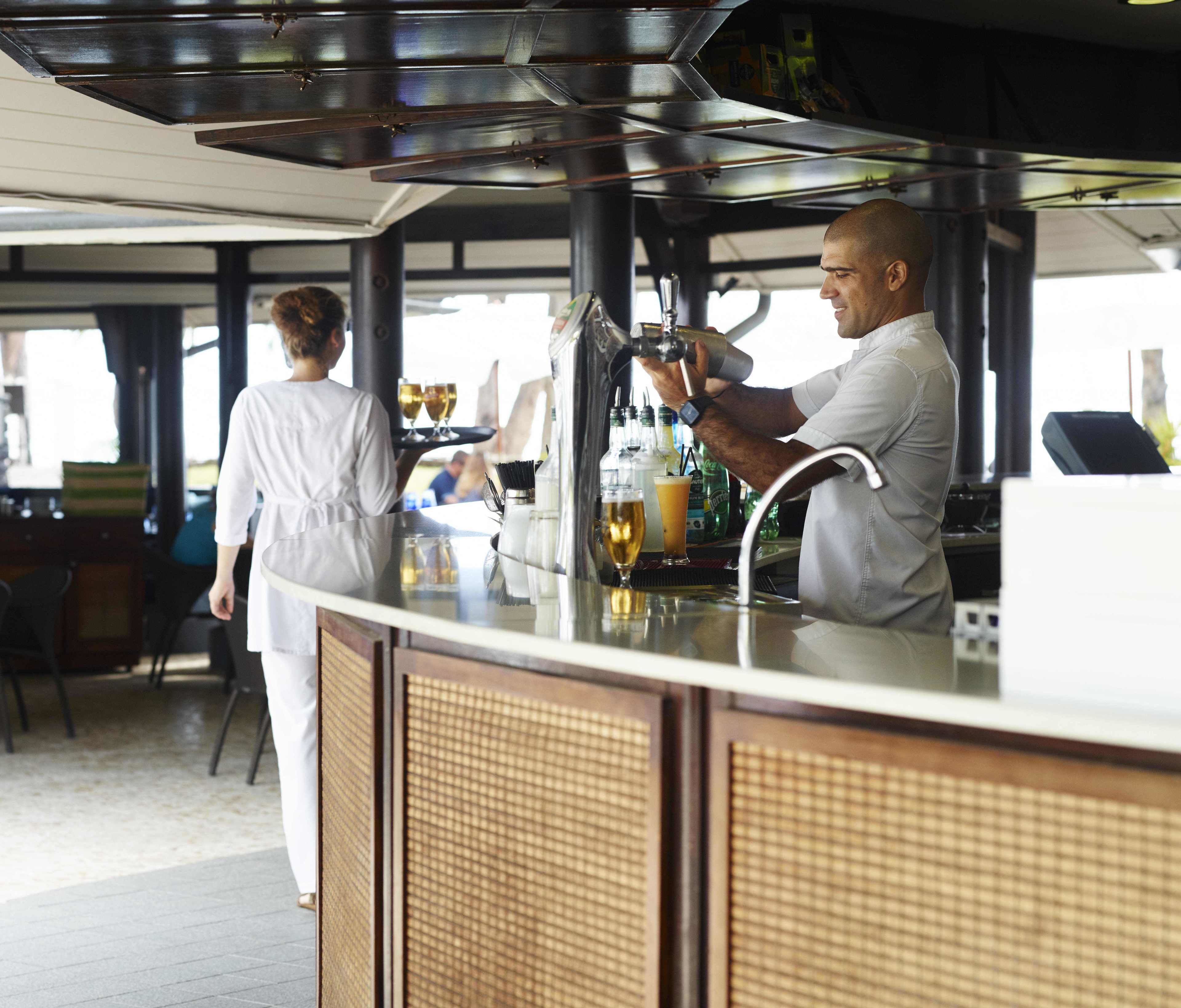 A bar area with a bartender preparing drinks. There are stools and a counter with a menu.