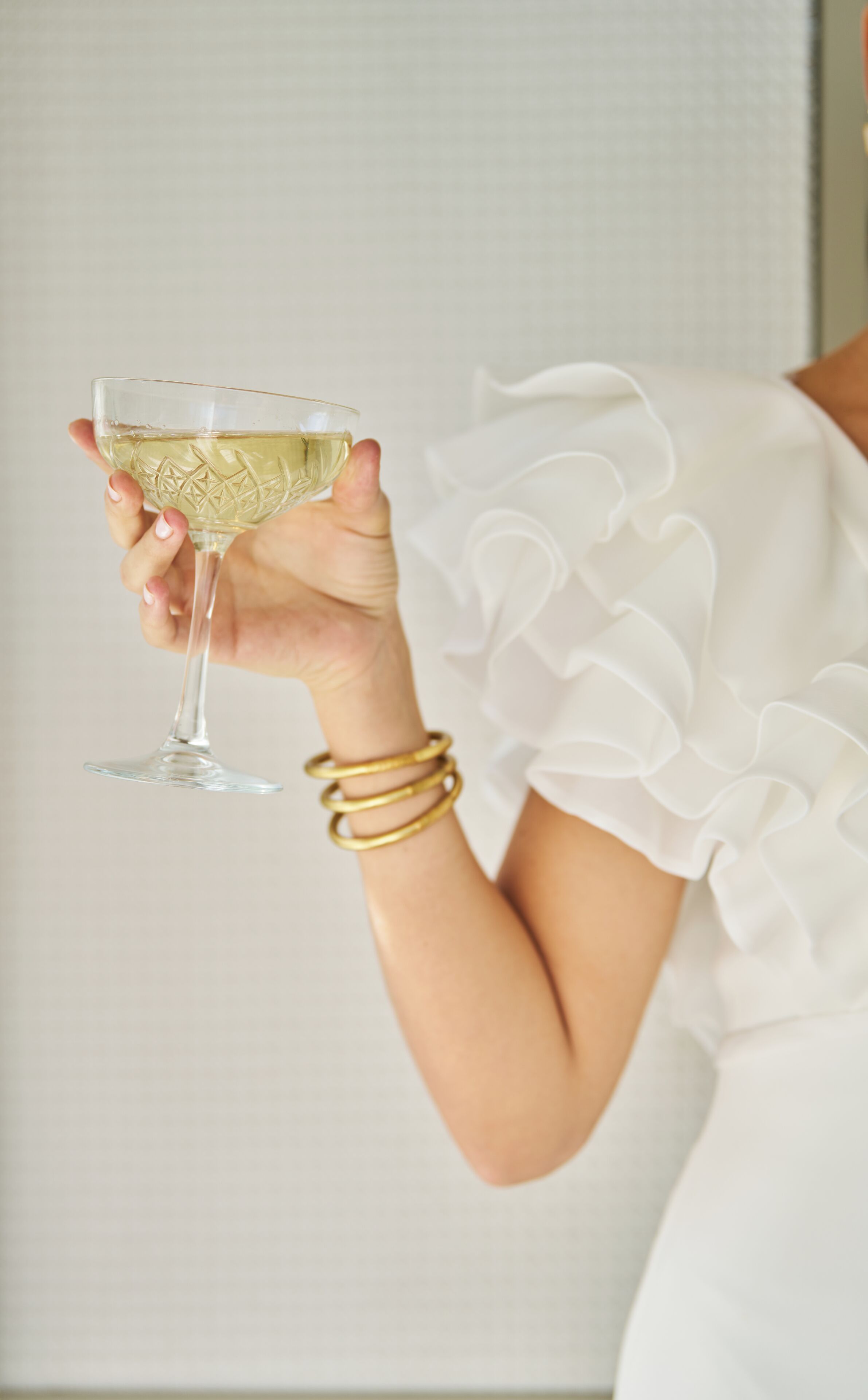 Una persona sosteniendo un vaso con líquido blanco, vestida con una prenda blanca de mangas fruncidas y brazaletes dorados.