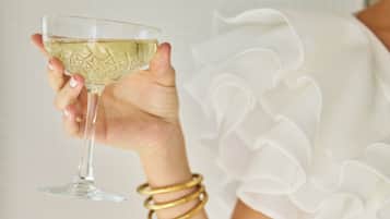 Una persona sosteniendo un vaso con líquido blanco, vestida con una prenda blanca de mangas fruncidas y brazaletes dorados.