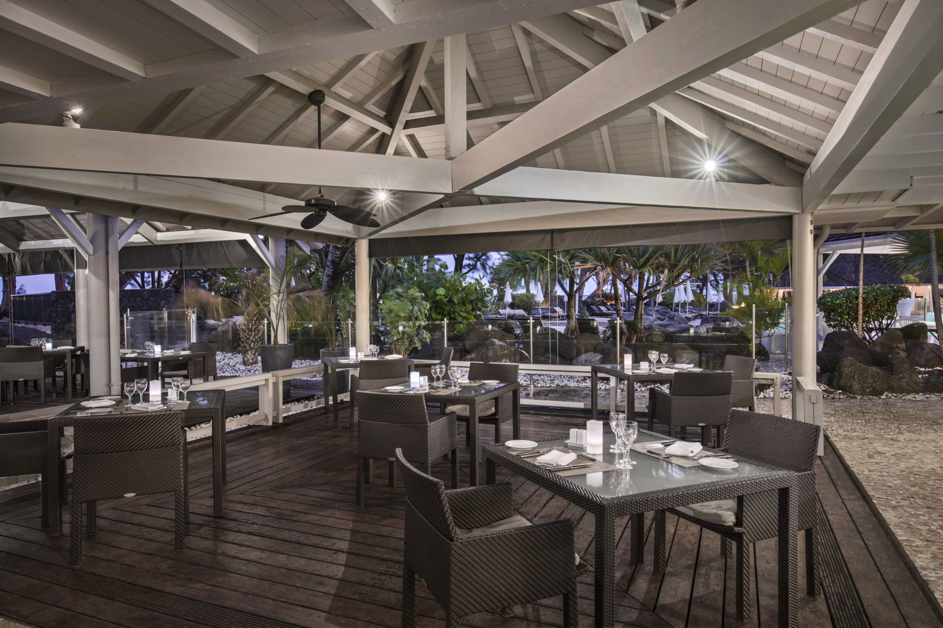 An outdoor dining area with wooden tables and chairs, a covered roof, and a view of palm trees and a pool.
