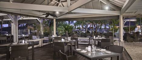 An outdoor dining area with wooden tables and chairs, a covered roof, and a view of palm trees and a pool.