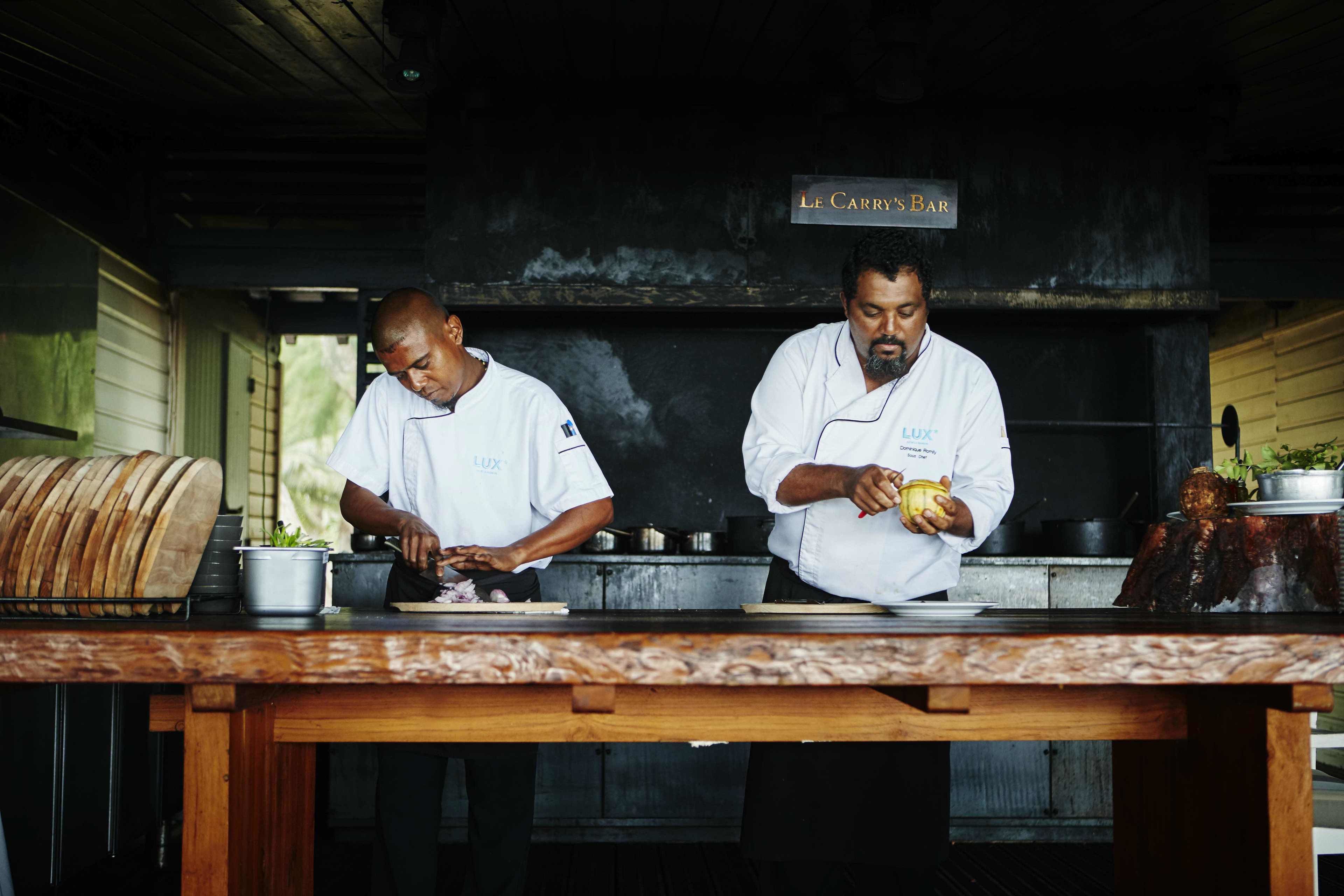 Two chefs preparing food at Le Carry's Bar, with a wooden table and kitchen equipment visible.