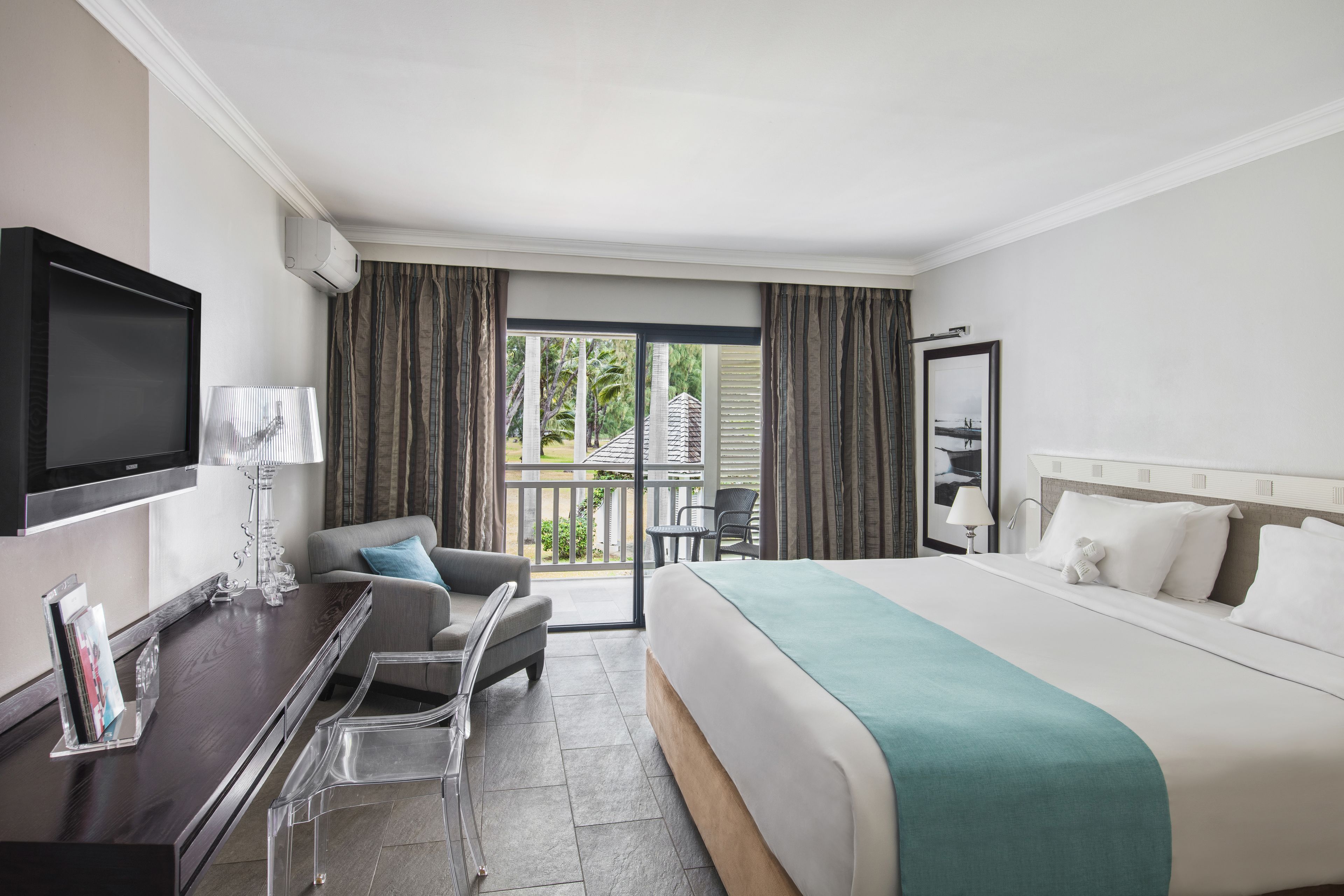 A hotel room with a bed, a desk with a TV, a chair, a balcony with curtains, and a view of greenery.