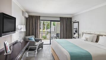 Habitación de hotel con cama, escritorio con televisor, silla, balcón con cortinas y vista a zonas verdes.