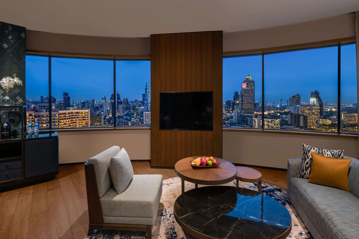 Una sala de estar moderna con un televisor grande, asientos cómodos, una mesita con fruta y vistas a la ciudad a través de amplios ventanales.