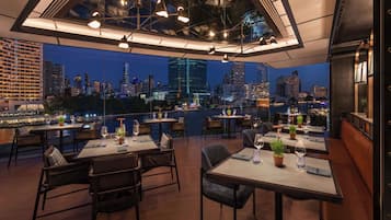 A rooftop restaurant with tables and chairs, city skyline views, and hanging lights.