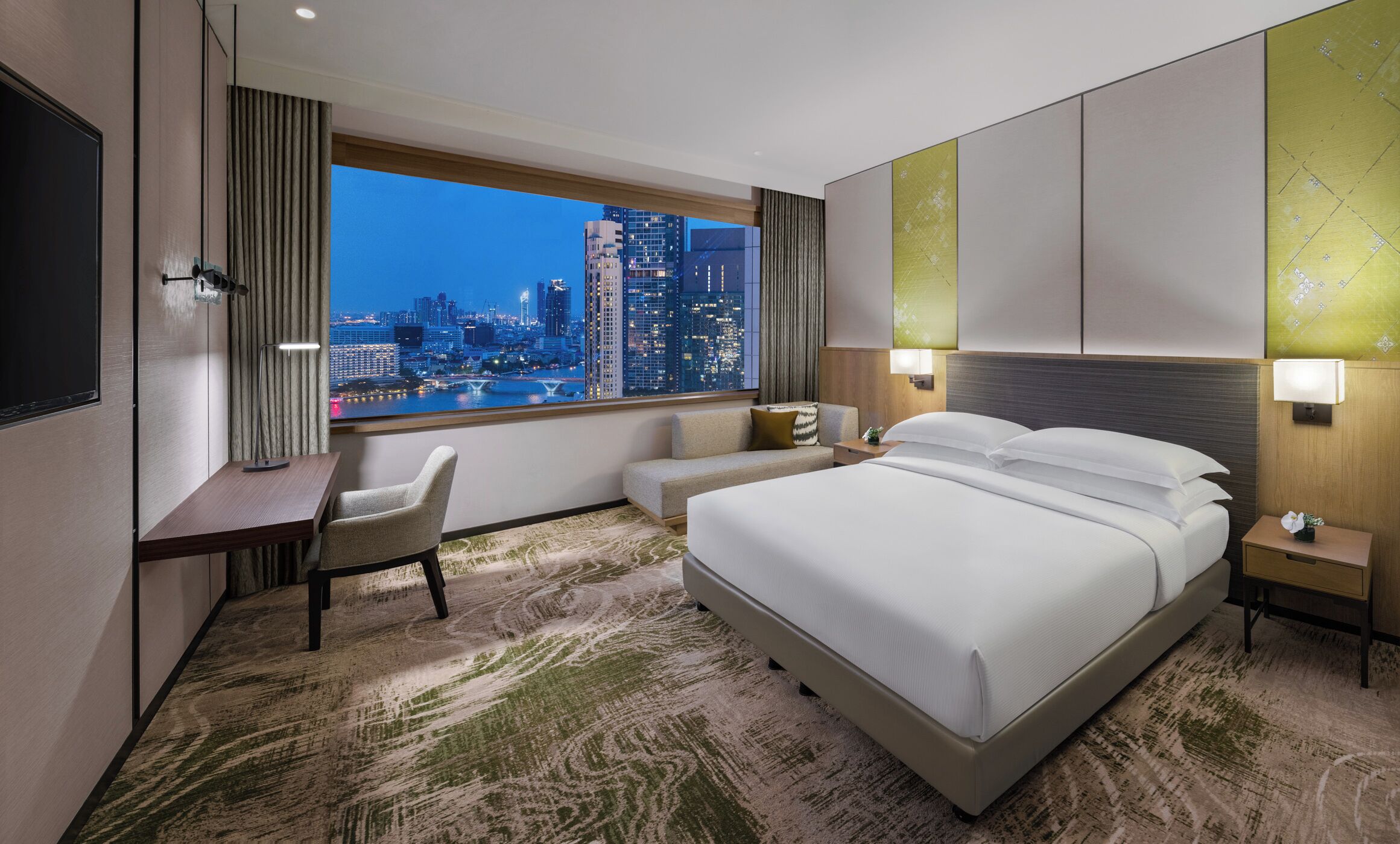 A modern hotel room with a large bed, a desk, and a view of a cityscape.
