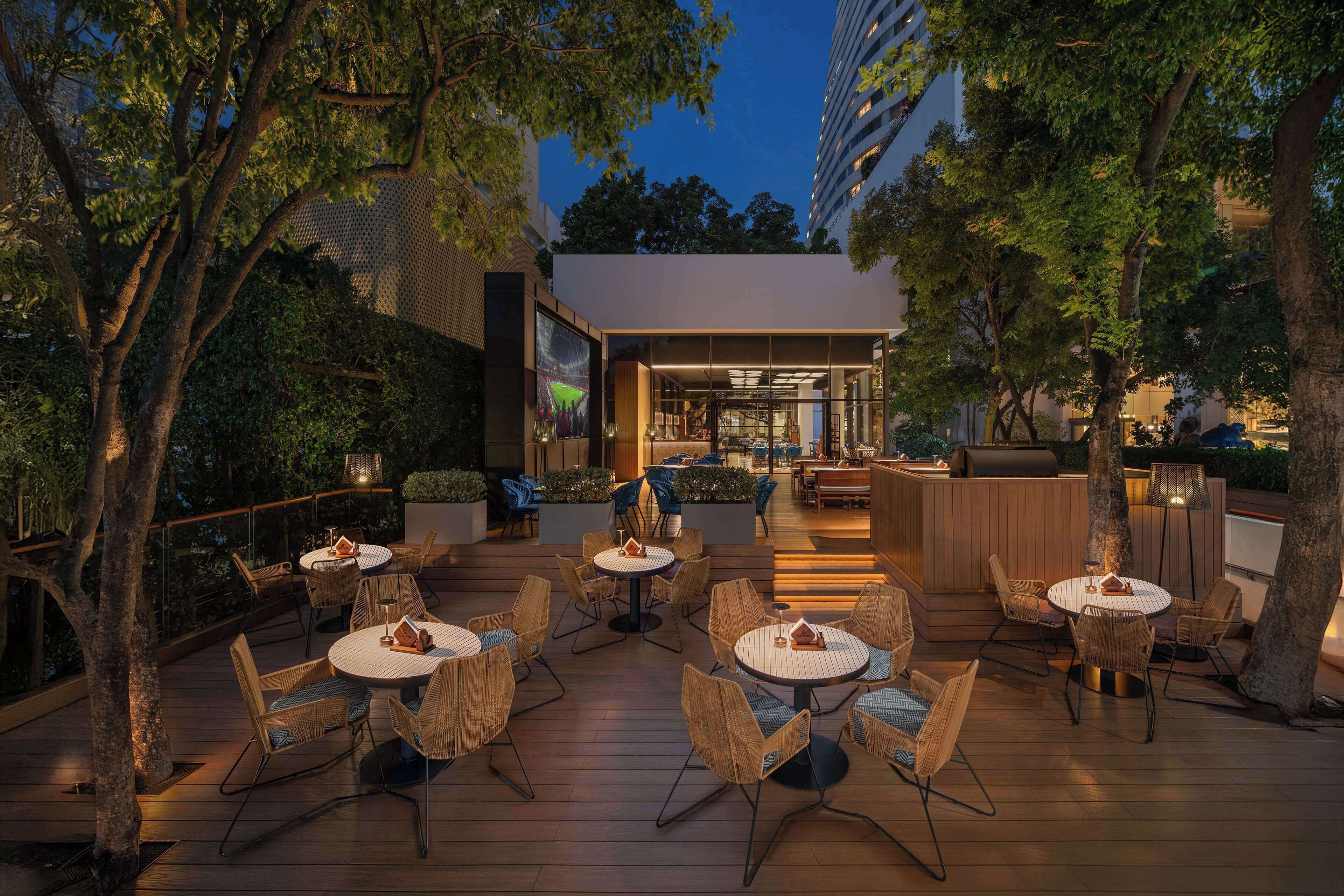 An outdoor patio area with round tables and wicker chairs, surrounded by trees and a building entrance.