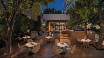 An outdoor patio area with round tables and wicker chairs, surrounded by trees and a building entrance.