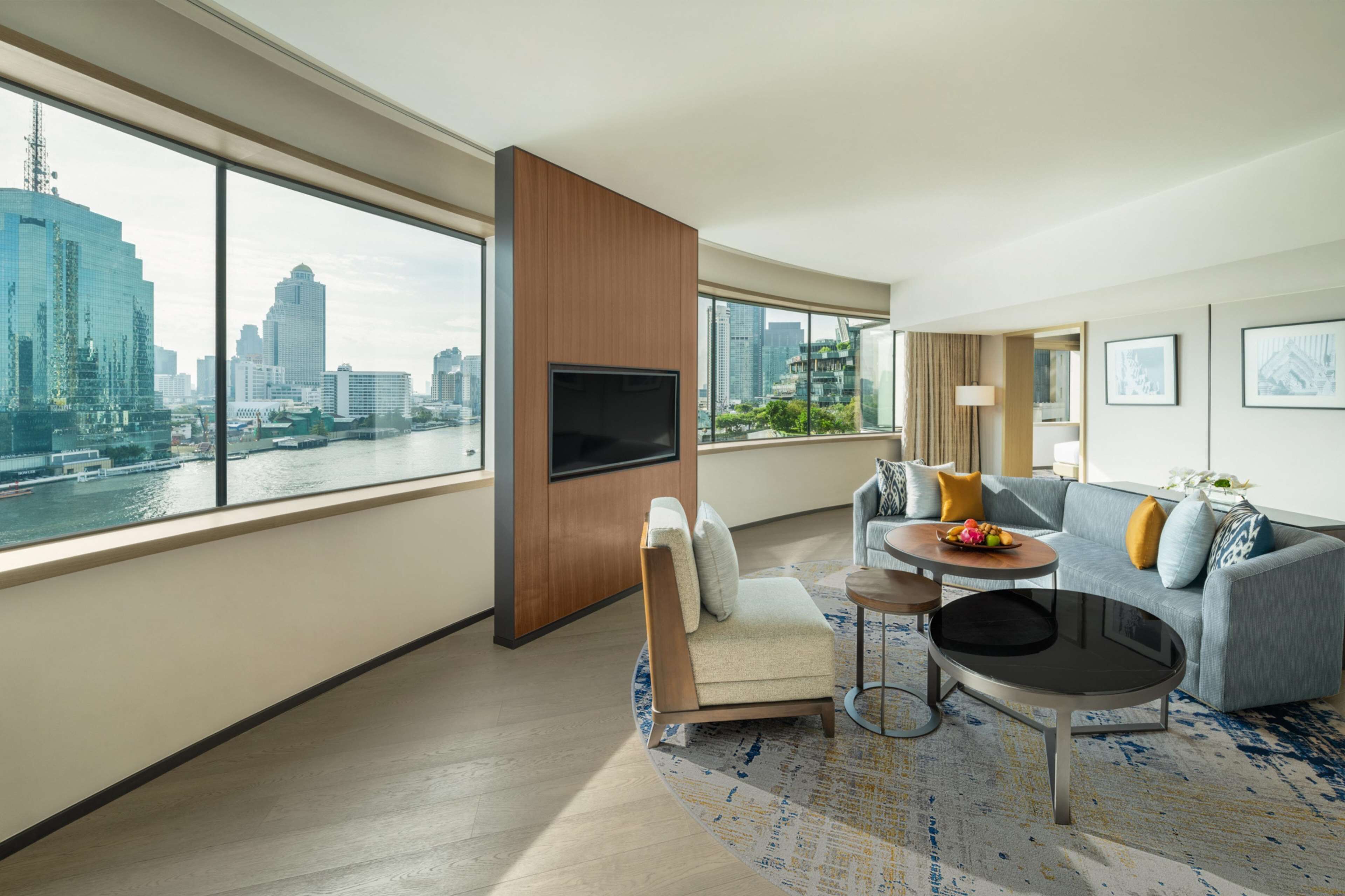 A modern hotel room with a large window offering a city view, a sofa, a small round table, and a TV mounted on a wooden panel.