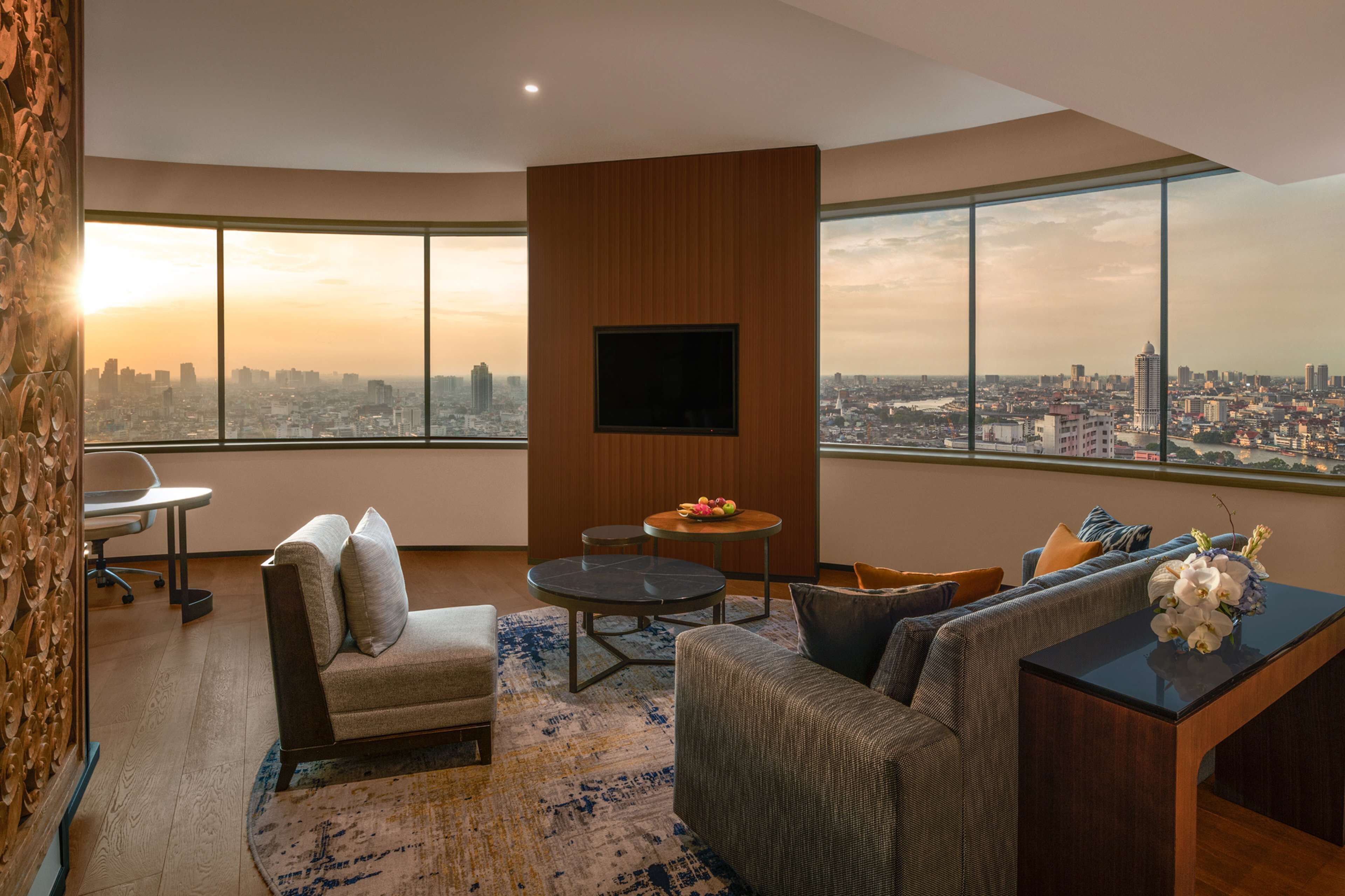 A modern living room with a city view, a flat-screen TV mounted on a wooden panel, a sofa, a coffee table, and a small round table with fruit.