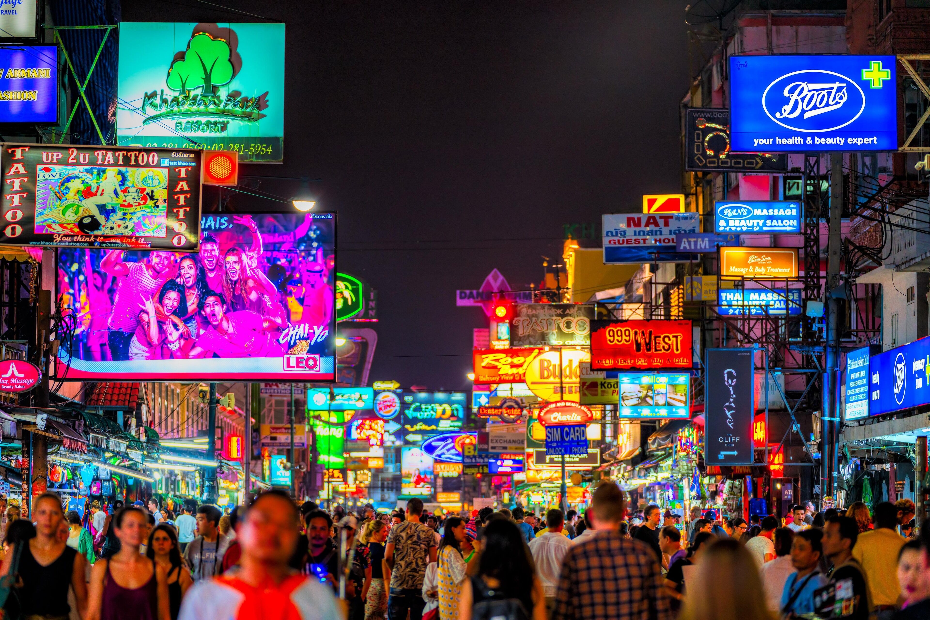 A busy street with neon signs and billboards, including a tattoo parlor and a massage salon.