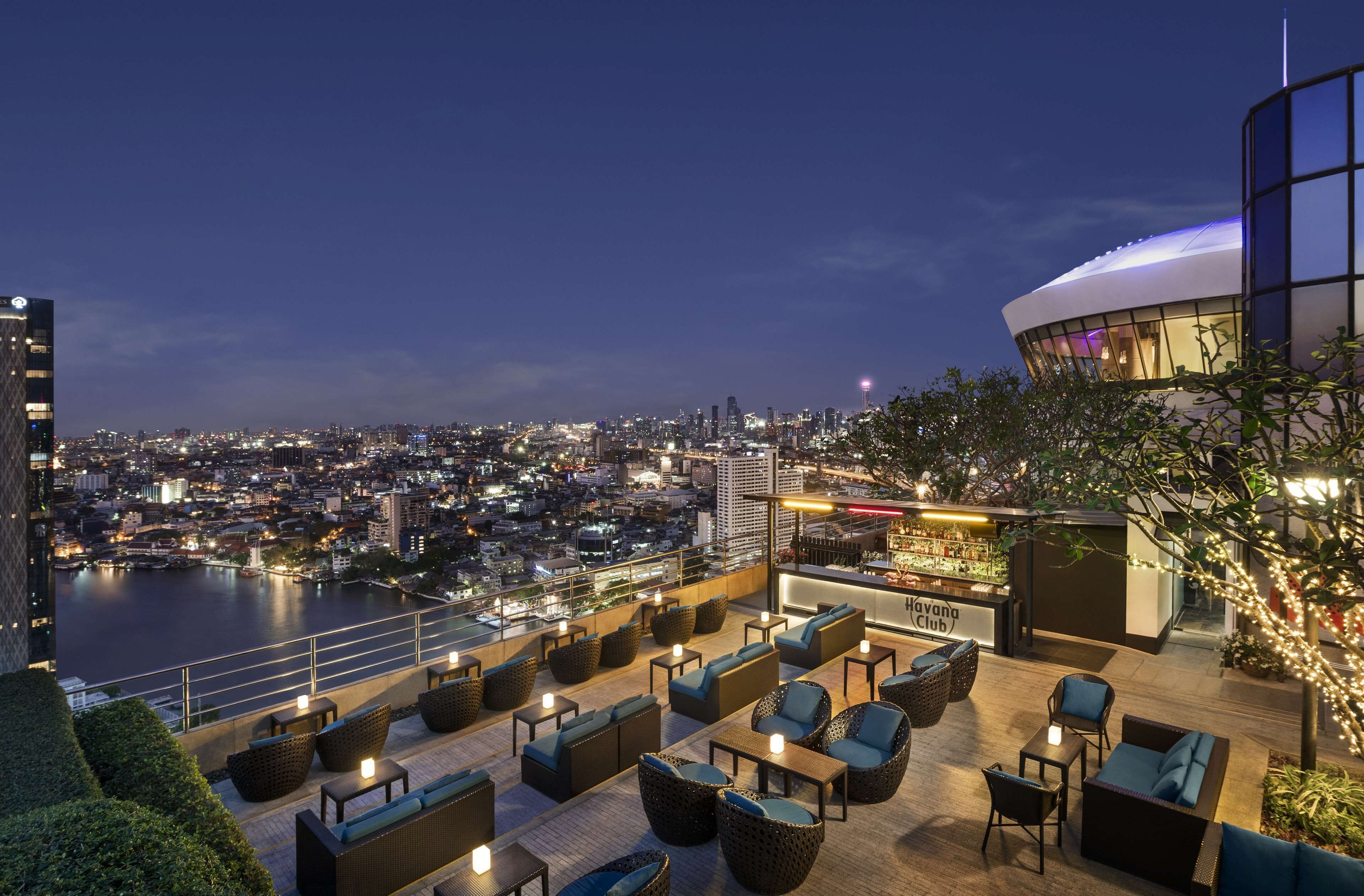 Un bar en la azotea con mesas al aire libre, vista panorámica de la ciudad y un edificio moderno con una estructura en forma de cúpula.