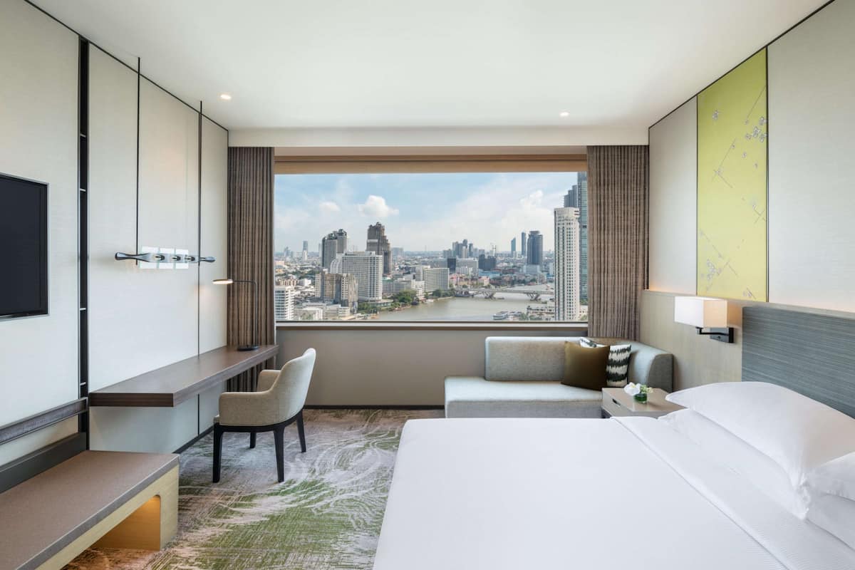 Habitación de hotel con una cama grande, un escritorio, una silla y vistas a un paisaje urbano a través de un amplio ventanal.
