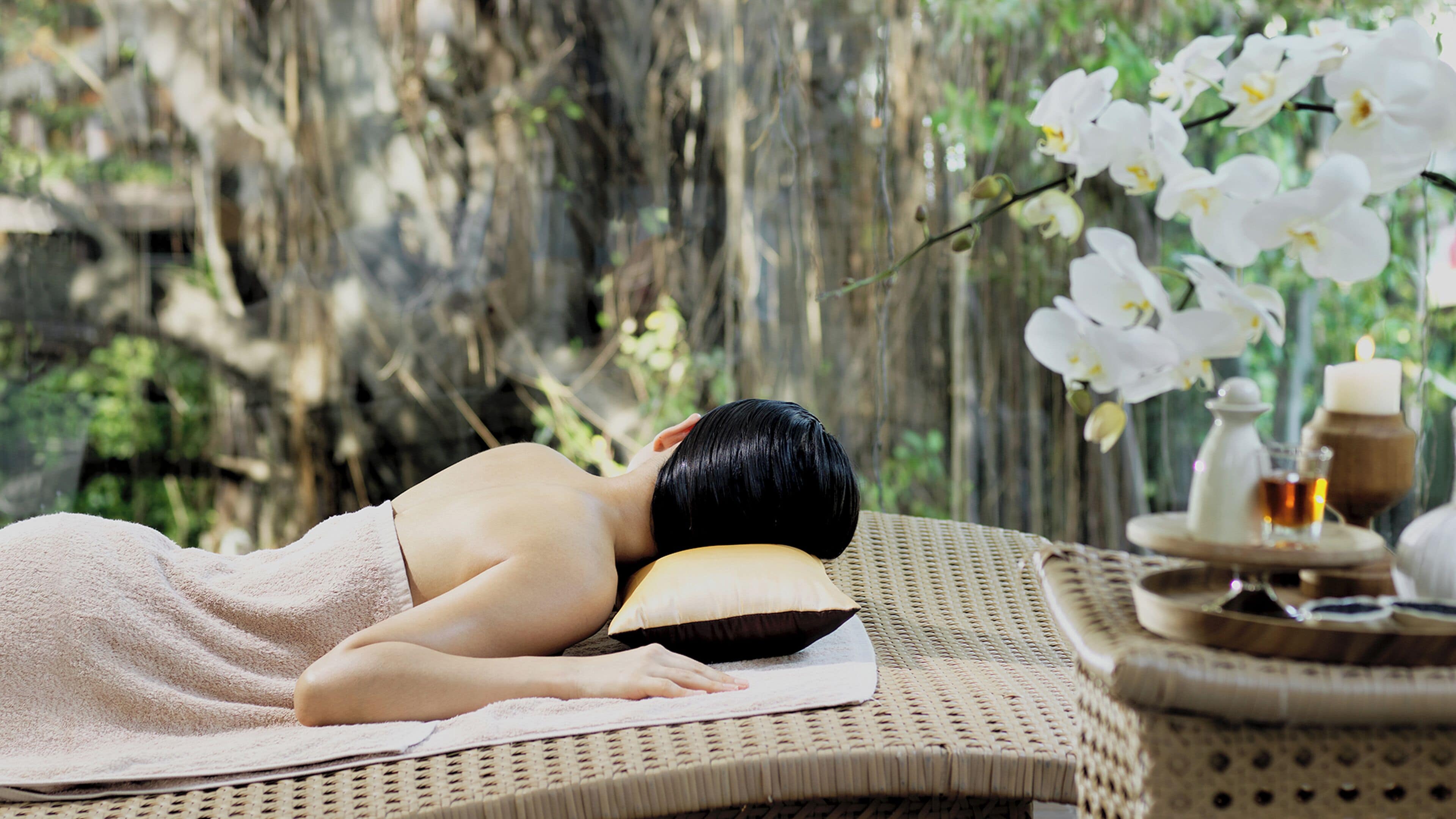 A person lying on a massage table with a towel, surrounded by a wicker chair, a candle, and a glass of water.