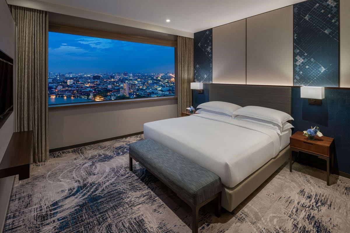 Habitación de hotel moderna con una cama grande, un banco, una mesita de noche y vistas a un paisaje urbano al atardecer.