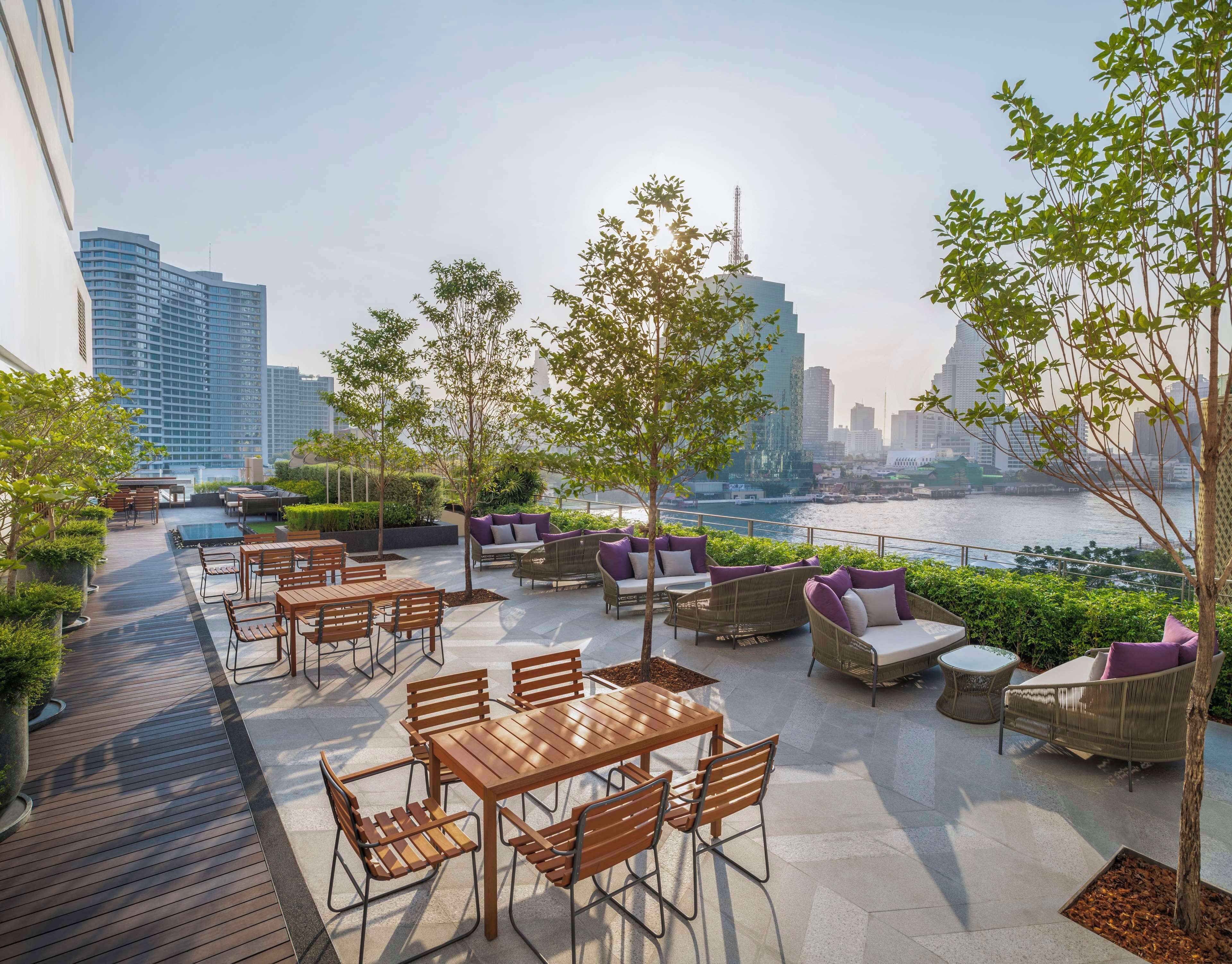 An outdoor seating area with tables and chairs, surrounded by greenery and overlooking a river with a city skyline in the background.