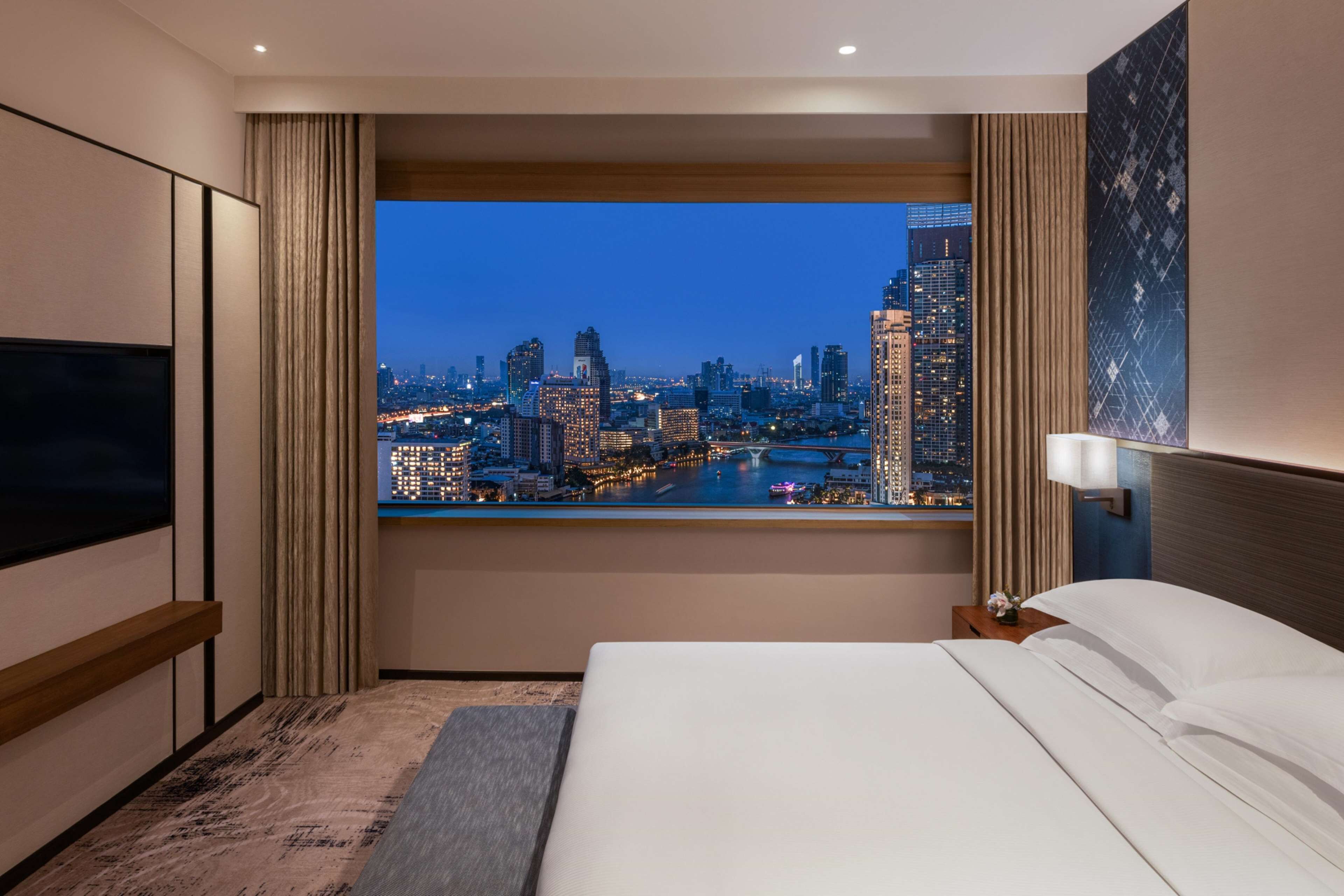 A hotel room with a large bed, a TV, and a city view through a window.