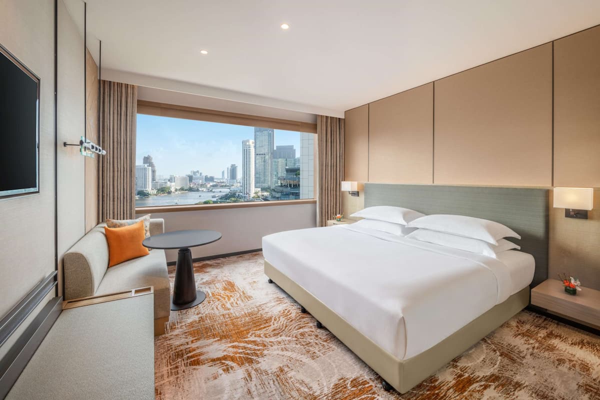 Habitación de hotel con una cama grande, un sofá, una mesita y vistas a un paisaje urbano a través de un ventanal.