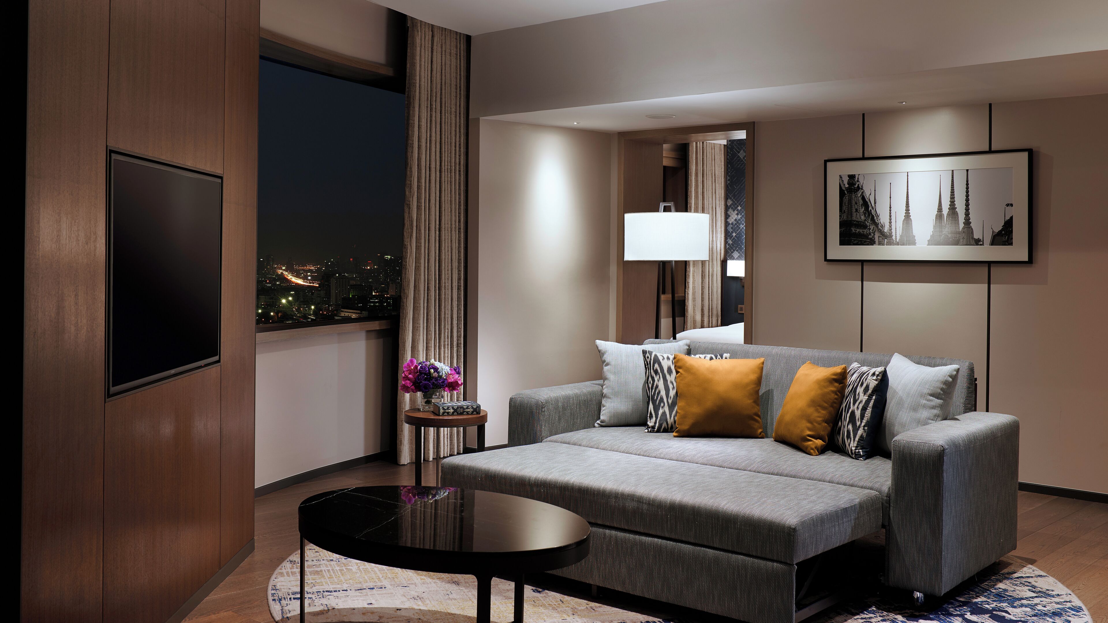 A modern hotel room with a grey sofa, a black coffee table, and a cityscape view through the window.