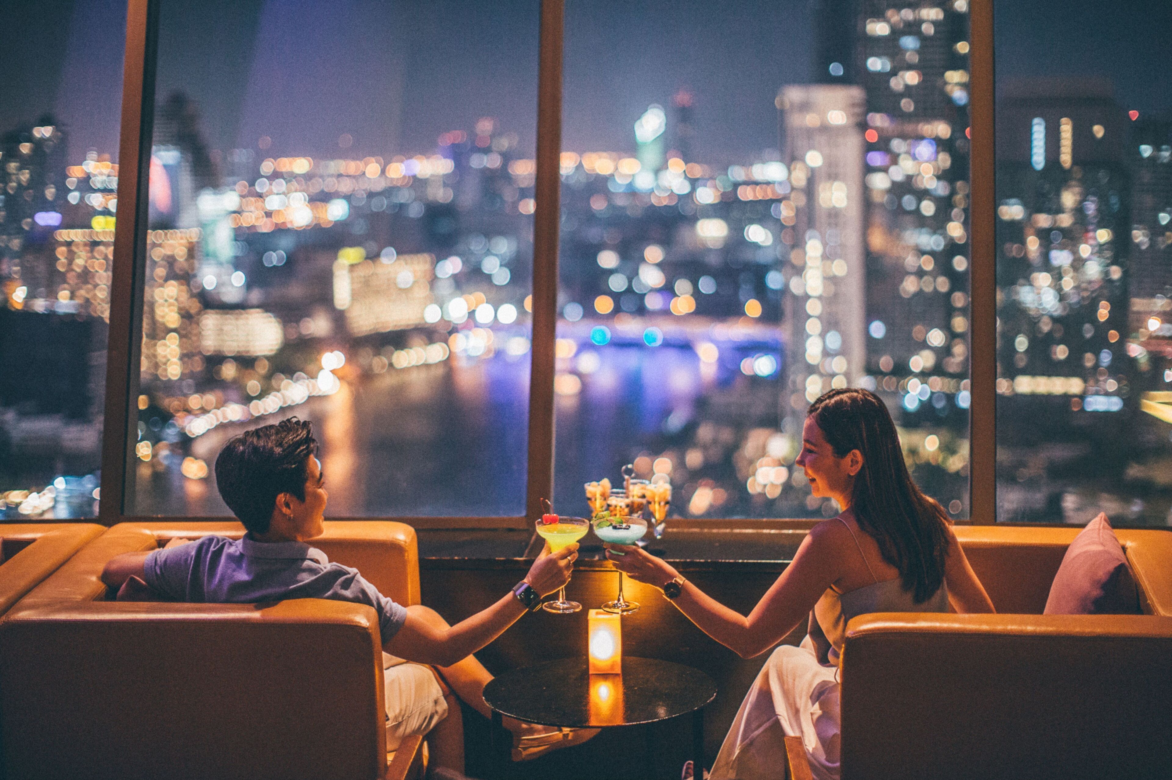 A couple seated at a table with drinks, overlooking a cityscape at night.