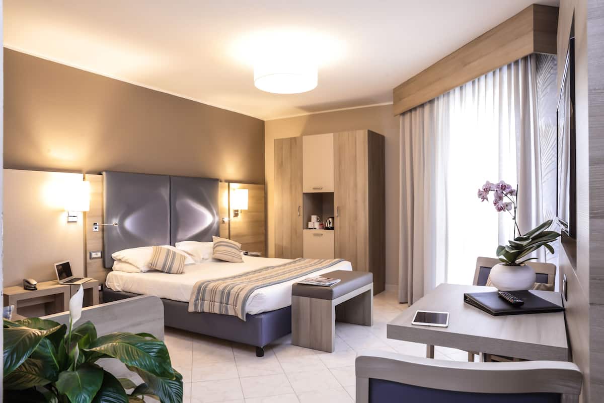 Ein modernes Hotelzimmer mit einem großen Bett, Nachttischen, einem Schreibtisch mit Stuhl und einer Zimmerpflanze.