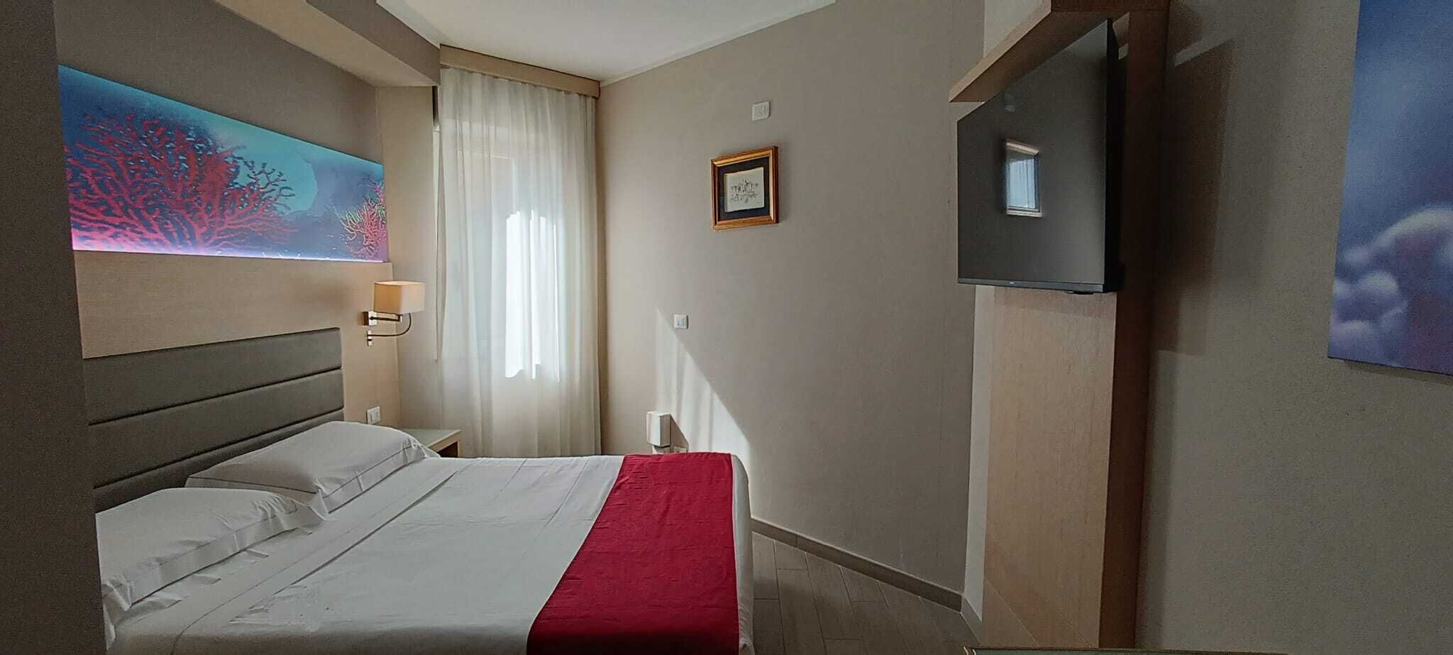Una camera d'albergo con un letto, una televisione, un quadro e una finestra con tende.