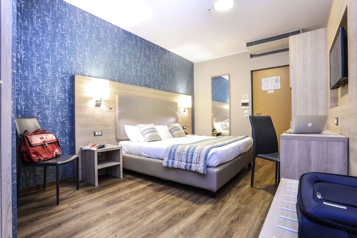 Ein modernes Hotelzimmer mit einem großen Bett, einem Schreibtisch mit einem Laptop, einem Sessel und einer roten Handtasche auf Holzdielen.