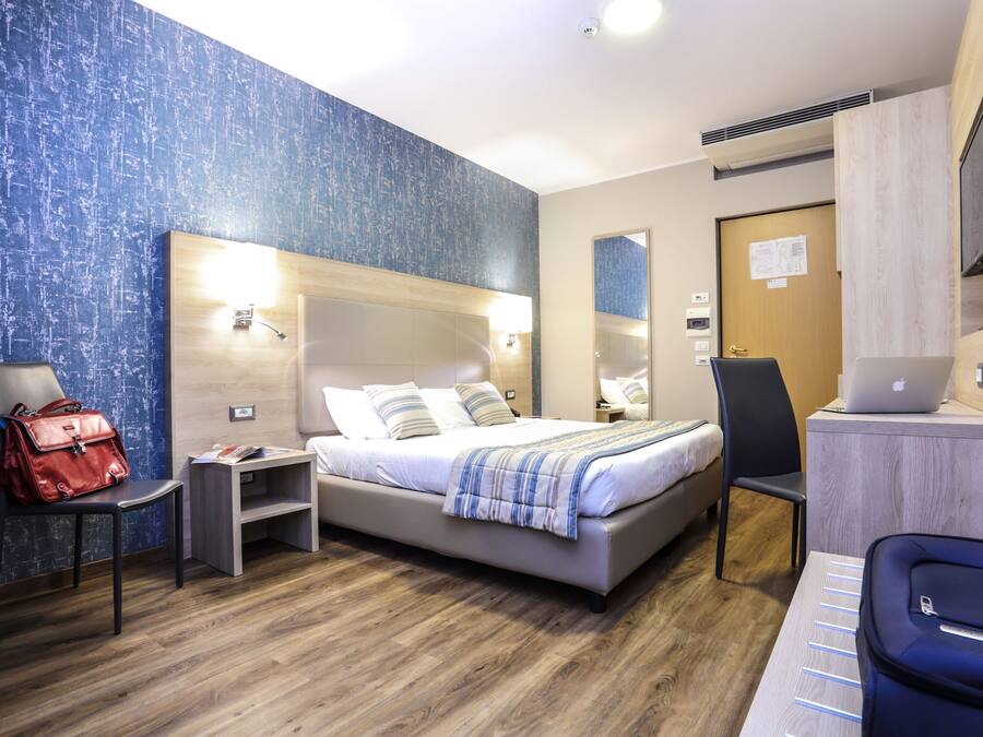 Quarto de hotel moderno com uma cama grande, uma escrivaninha com um laptop, uma cadeira, uma bolsa vermelha e piso de madeira.