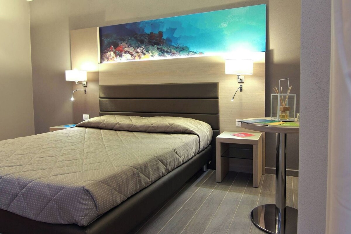 A hotel room with a bed, bedside lamps, a small table with a lamp, and a painting on the wall.