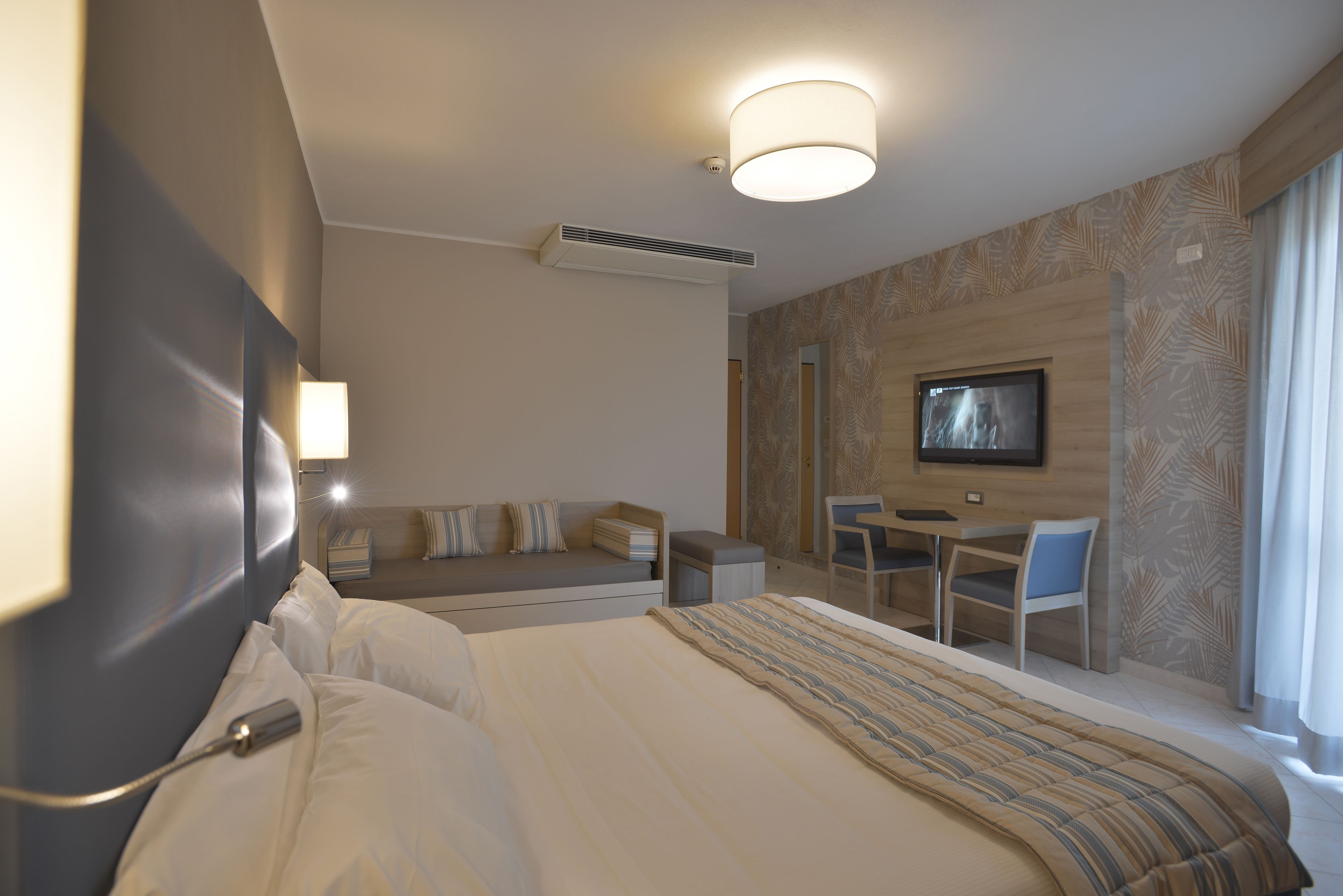 Quarto de hotel com uma cama grande, uma escrivaninha com cadeira, televisão e um sofá.