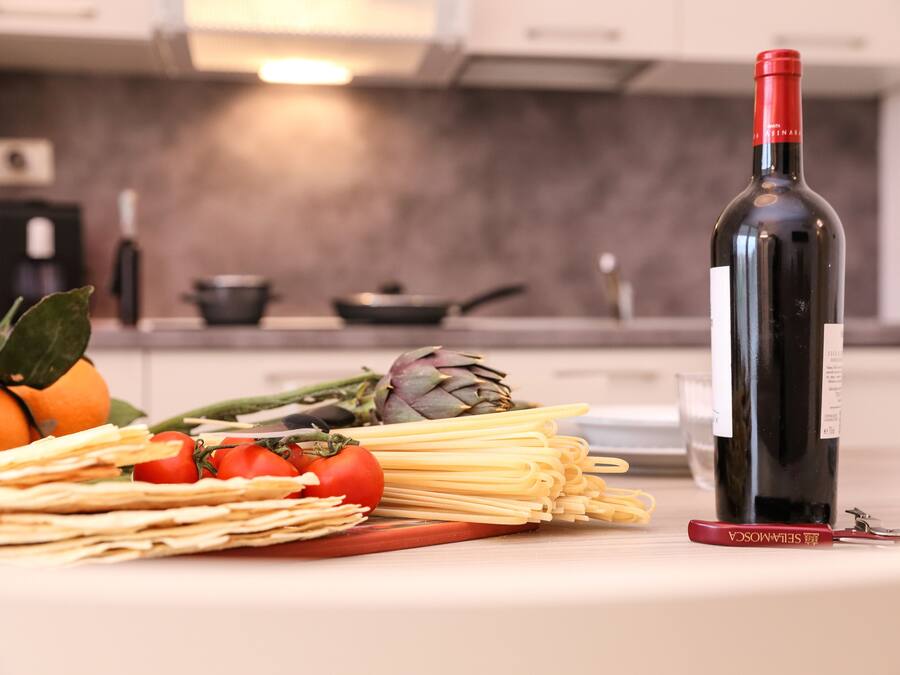 Una encimera de cocina con una botella de vino tinto, pasta, tomates, alcachofas y un sacacorchos.