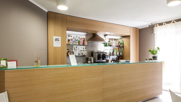 A bar area with a wooden counter, shelves of liquor bottles, a coffee machine, and a potted plant. - Alma Di Alghero Hotel (Alghero)