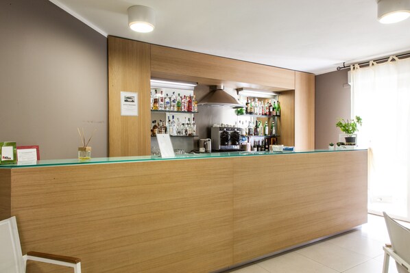 A bar area with a wooden counter, shelves of liquor bottles, a coffee machine, and a potted plant. - Alma Di Alghero Hotel (Alghero)