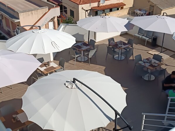An outdoor seating area with multiple white umbrellas and tables, surrounded by buildings. - Alma Di Alghero Hotel (Alghero)