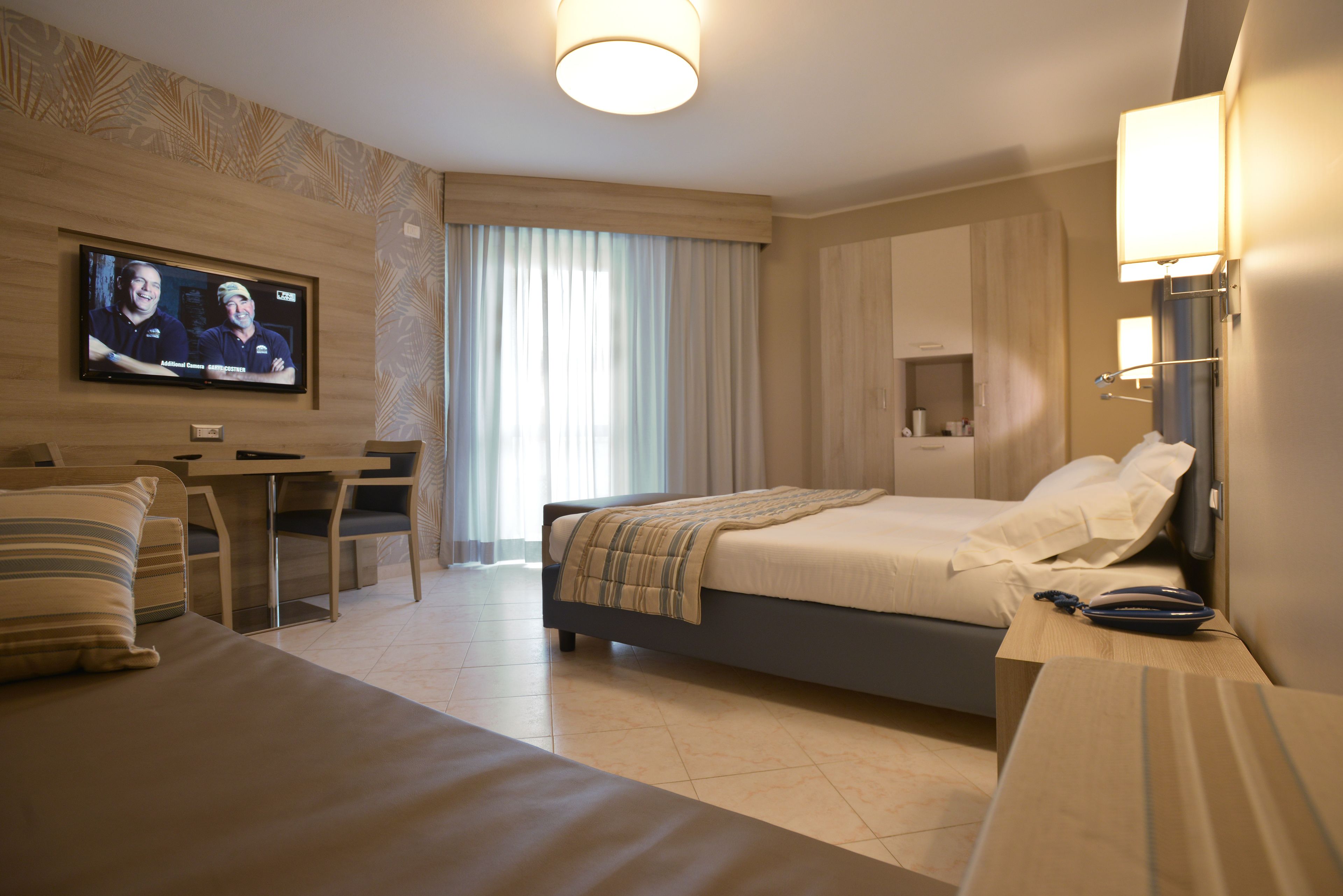 A modern hotel room with a bed, a desk, a television, and a large window with curtains.