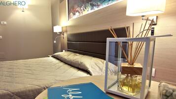 Una cama bien hecha con cubrecolchón acolchado, una mesita de noche con un libro y un recipiente de vidrio con ramitas de junco, y una lámpara de pared.