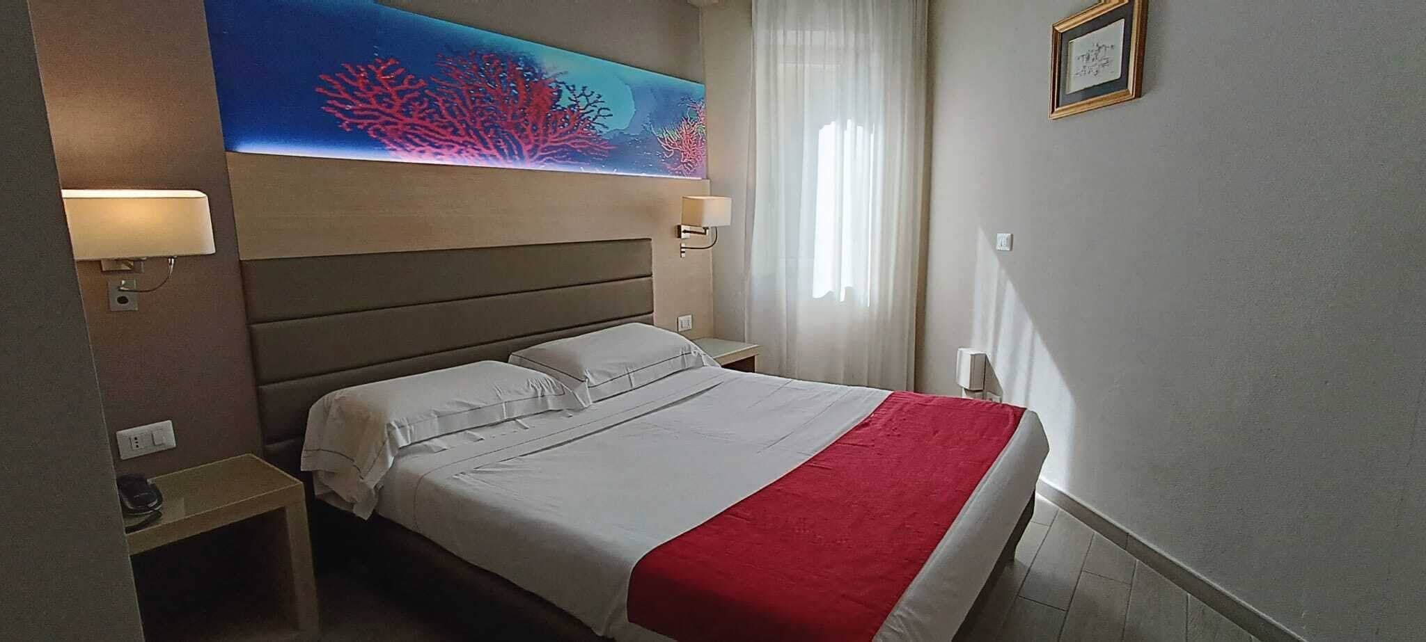 Habitació d'hotel amb llit, taulell, làmpada, quadre i finestra amb cortines.