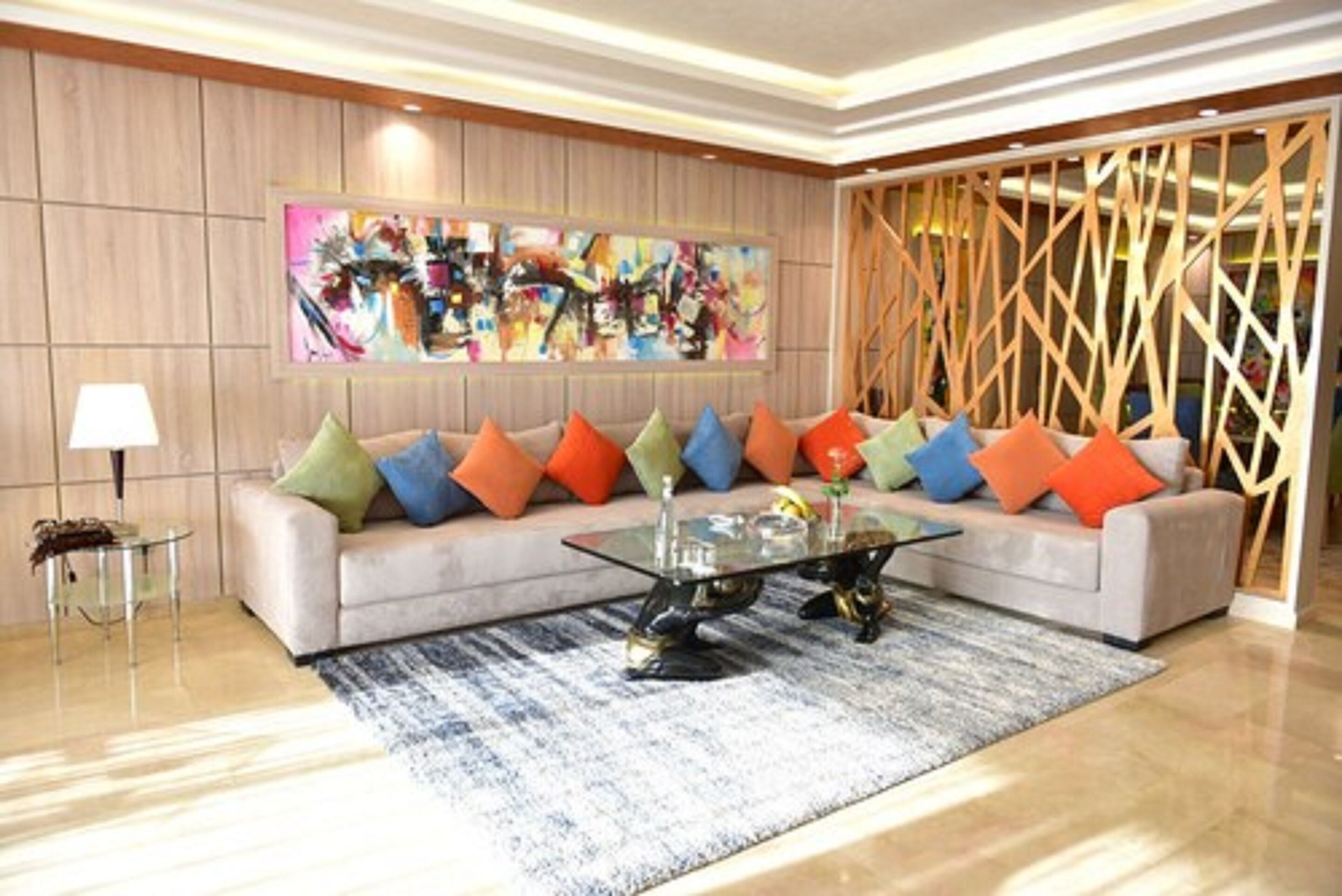 Una sala de estar moderna con un sofá color crema, cojines coloridos, una mesa de centro de vidrio y un cuadro abstracto grande en la pared.