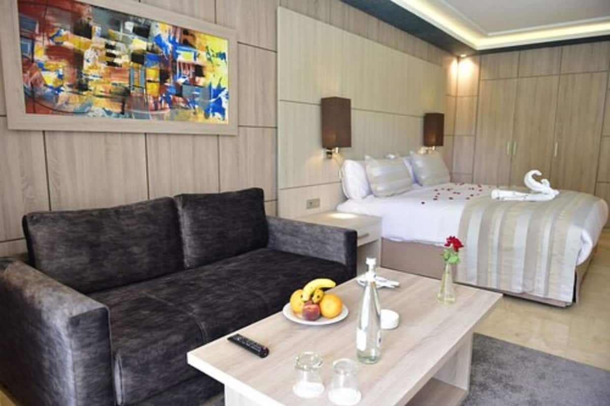 A modern hotel room with a grey sofa, a bed with white bedding, a painting on the wall, and a small table with a fruit plate and a vase.