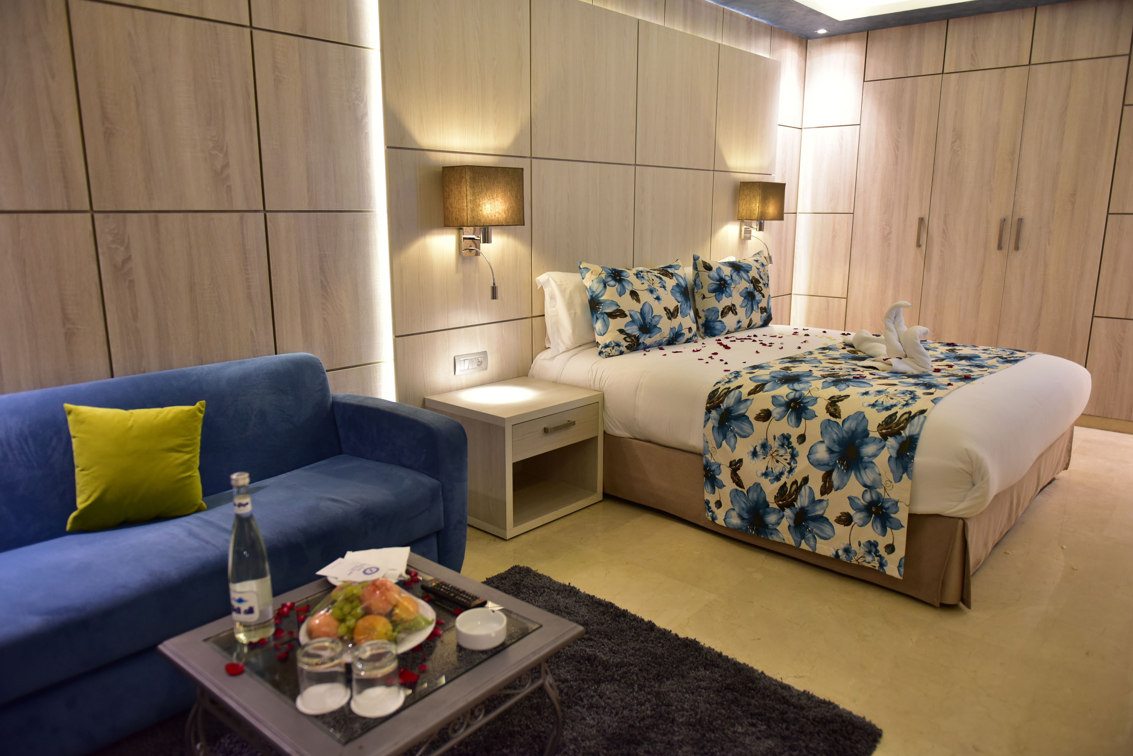 A hotel room with a bed, a blue sofa, a nightstand, and a coffee table with a bottle and glasses.