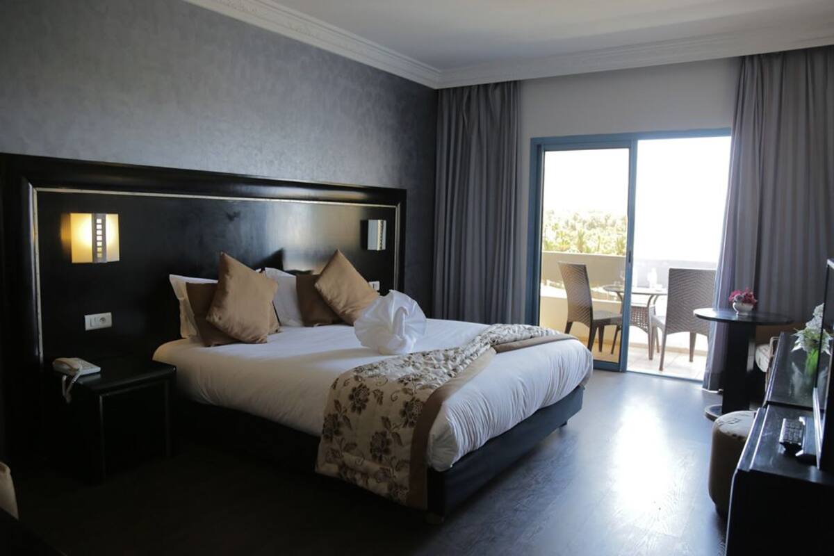 Habitación de hotel con una cama grande, una mesita de noche con lámpara, una silla, una pequeña mesa con un florero y vistas al exterior a través de una puerta corredera.
