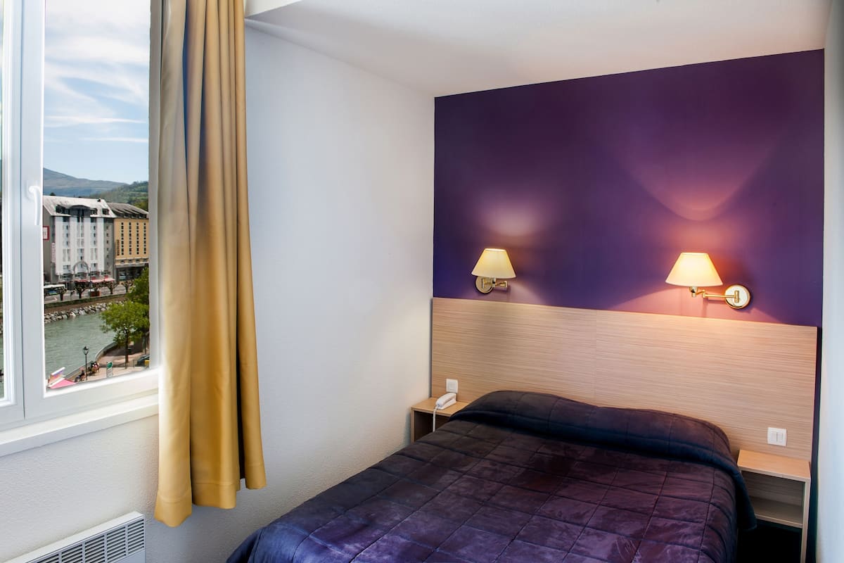 A hotel room with a bed, a nightstand, a lamp, and a view of a cityscape through a window with curtains.