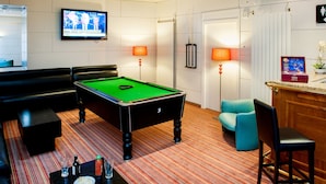 A pool table with a green felt surface, a billiards cue, and a black pool table with a number 3 on it. There are two lamps with orange shades, a television mounted on the wall, and a bar area with a countertop and a potted plant. - Hotel Continental (Lourdes)