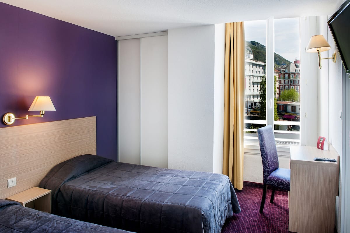 A hotel room with two beds, a desk, a chair, a lamp, and a window with a view of buildings.